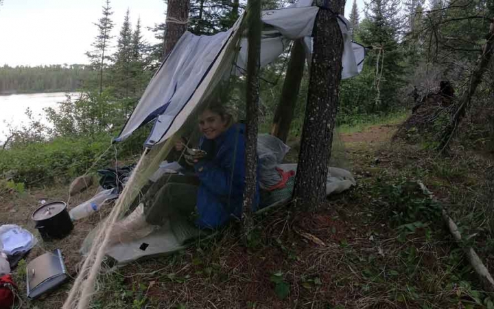a student smiles from a tarp shelter in a wooded area beside a body of water 